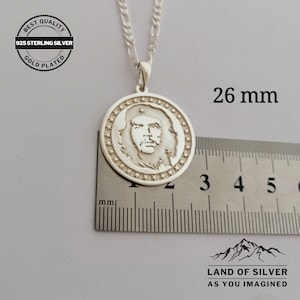 May include: Silver pendant necklace featuring a round, gold-plated charm with a profile image of a man. The charm is 26 mm in diameter and has a star-shaped border. The necklace is on a silver chain.