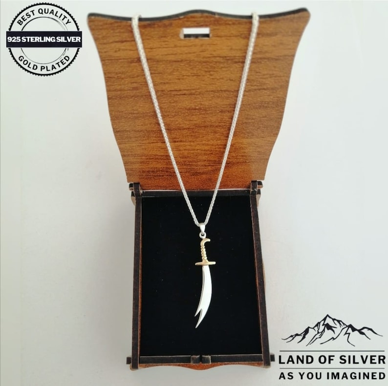 May include: A silver chain necklace with a gold-plated pendant shaped like a scimitar sword. The necklace is in a wooden box with a black velvet lining. The box has a label that reads "Land of Silver as you imagined".