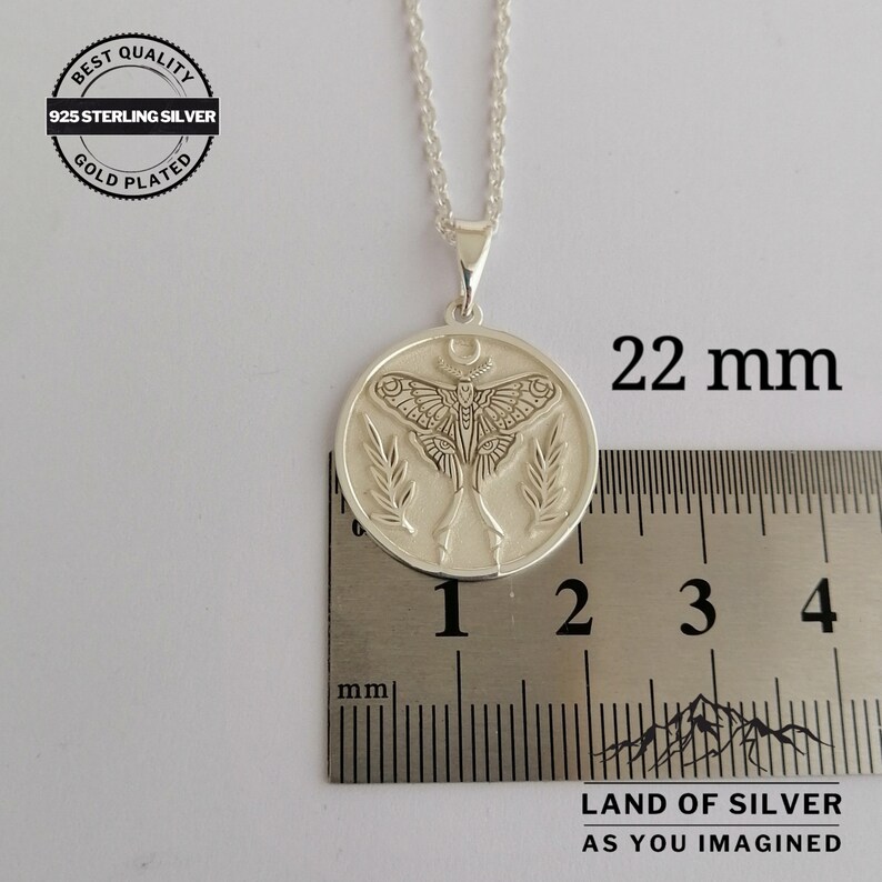 May include: A silver pendant necklace with a detailed moth design, measuring 2.2 cm in diameter. The pendant features a moth with intricate wing patterns, a crescent moon, and leaf accents. The necklace is labeled "925 Sterling Silver" and "Gold Plated".