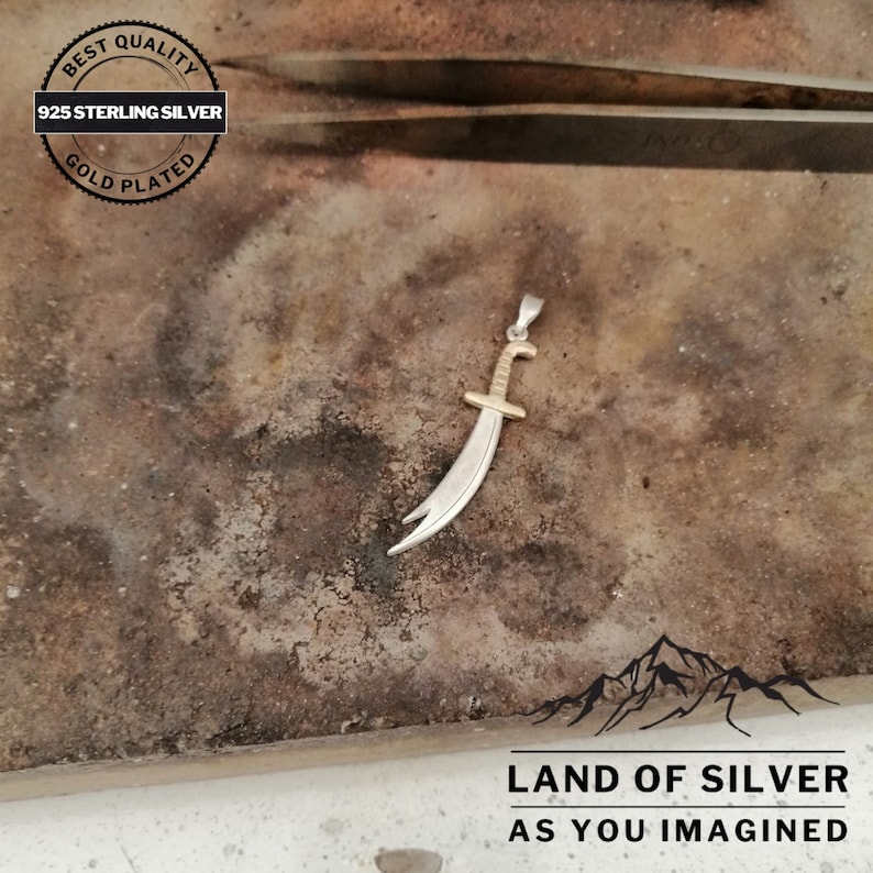 May include: A silver pendant in the shape of a scimitar sword with gold accents. The pendant is on a rough, brown surface. The text "LAND OF SILVER AS YOU IMAGINED" is visible below the pendant.