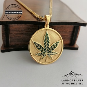 May include: A gold plated 925 sterling silver pendant necklace featuring a green marijuana leaf design. The pendant is round and hangs from a gold chain. The necklace is displayed in a wooden box with the words "Land of Silver as you imagined" on the bottom.
