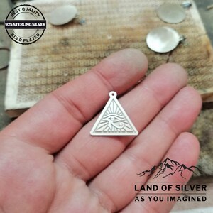 May include: Silver pendant charm in the shape of a triangle with an eye inside. The eye is surrounded by rays of light. The pendant is made of 925 sterling silver and is gold plated. The pendant is on a white surface.