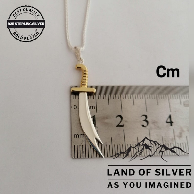 May include: A silver and gold pendant necklace featuring a stylized sword design. The pendant is approximately 4 cm long and is measured against a ruler. The necklace is labeled "925 Sterling Silver, Gold Plated" and "Land of Silver, As You Imagined".