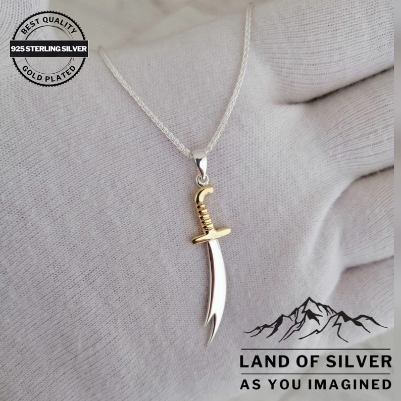 May include: A silver chain necklace with a gold-plated pendant shaped like a scimitar sword. The pendant has a silver blade and a gold handle. The necklace is on a white surface with the text "LAND OF SILVER AS YOU IMAGINED" below the pendant.