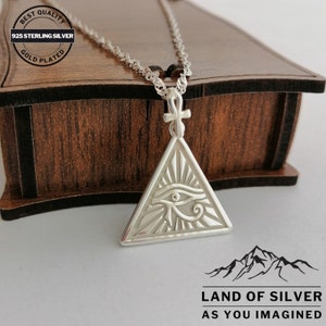 May include: Silver necklace with an eye of Horus pendant in a triangle shape. The pendant is detailed with an ankh symbol at the top and rays of light emanating from the eye. The necklace is on a wooden box with a label that reads "Best Quality 925 Sterling Silver Gold Plated".