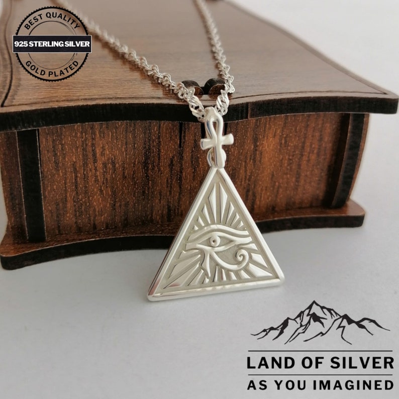 May include: Silver necklace with an eye of Horus pendant in a triangle shape. The pendant is detailed with an ankh symbol at the top and rays of light emanating from the eye. The necklace is on a wooden box with a label that reads "Best Quality 925 Sterling Silver Gold Plated".