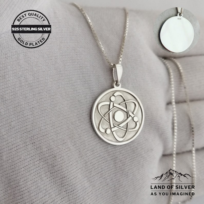 May include: A silver necklace with a round pendant featuring a stylized atom design. The pendant is 925 sterling silver and gold plated. The necklace is on a white surface with the text "LAND OF SILVER AS YOU IMAGINED" below the pendant.