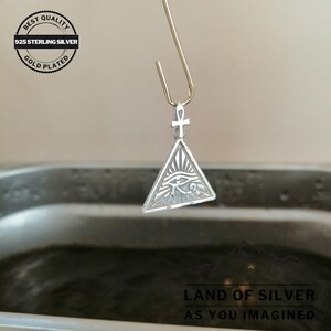 May include: A silver pendant with an eye of Horus design inside a triangle, with an ankh symbol at the top. The pendant is hanging from a gold-colored hook over a dark liquid.