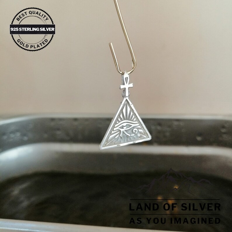 May include: A silver pendant with an eye of Horus design inside a triangle, with an ankh symbol at the top. The pendant is hanging from a gold-colored hook over a dark liquid.
