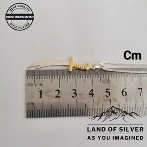 May include: A silver and gold pendant in the shape of a scimitar sword, measuring 4 cm in length, on a silver chain. The pendant is made of 925 sterling silver and is gold plated. The image includes the text "BEST QUALITY 925 STERLING SILVER GOLD PLATED" and "LAND OF SILVER AS YOU IMAGINED".