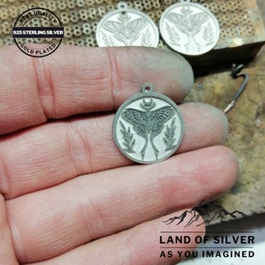 May include: A silver-coloured round pendant featuring a moth design, a crescent moon, and leaf accents. The pendant is stamped with "925 STERLING SILVER" and "GOLD PLATED". The image also includes the text "LAND OF SILVER AS YOU IMAGINED".