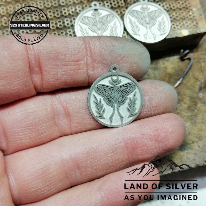 May include: A silver-coloured round pendant featuring a moth design, a crescent moon, and leaf accents. The pendant is stamped with "925 STERLING SILVER" and "GOLD PLATED". The image also includes the text "LAND OF SILVER AS YOU IMAGINED".