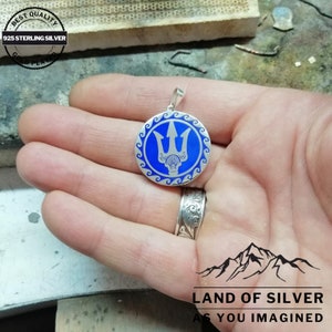 May include: A silver pendant with a blue enamel design of a trident, a symbol of Poseidon, the Greek god of the sea. The pendant is surrounded by a wave pattern. The pendant is made of 925 sterling silver and is gold plated.