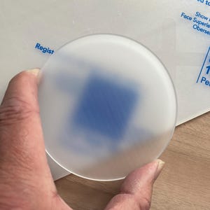 May include: A circular, frosted object with a blue square visible through it. The object is held by a hand. The background includes white paper with blue text that reads "Regist" and "PERSPE".