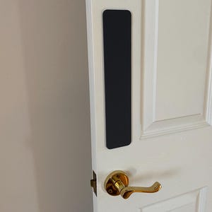 May include: A black door handle protector is attached to a white door. The door features a gold-coloured handle and a rectangular black pad to prevent scratches. The door is part of a white panelled door.