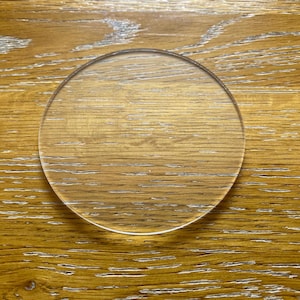 May include: A transparent, circular glass object, such as a coaster or lens, rests on a wooden surface. The glass is clear, revealing the wood grain beneath. The wood has a warm, golden-brown colour.