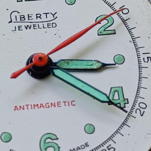 May include: Close-up of a vintage watch face. The white face features the word "LIBERTY JEWELLED" and "ANTIMAGNETIC" in red. The hands are red and turquoise, with the numbers 2 and 4 in green.