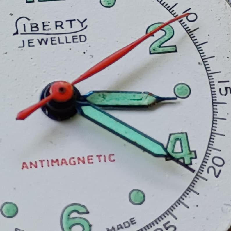 May include: Close-up of a vintage watch face. The white face features the word "LIBERTY JEWELLED" and "ANTIMAGNETIC" in red. The hands are red and turquoise, with the numbers 2 and 4 in green.