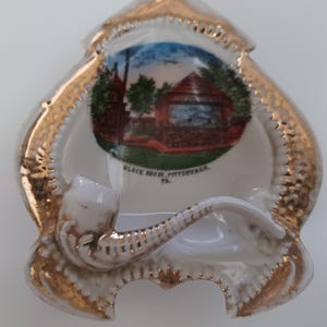 May include: A white ceramic ashtray with a gold-trimmed spade shape, featuring a central illustration of the Block House in Pittsburgh, PA. A white and gold ceramic pipe is attached to the front, serving as a rest for the ashtray.