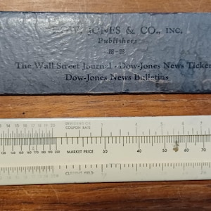 May include: An antique slide rule and a blue-grey paper sleeve with text from Dow Jones & Co., Inc. The slide rule is white with black markings and numbers. The paper sleeve has text including "The Wall Street Journal".