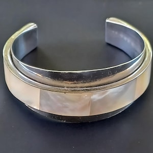 May include: A silver cuff bracelet with a rectangular mother-of-pearl inlay. The bracelet has a polished finish and a simple, elegant design. The bracelet is approximately 2.5 inches wide.