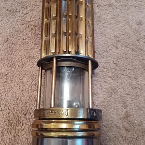 May include: A brass and silver miners lamp with a glass lens and a handle. The lamp has the words "Made in Germany" inscribed on the side.