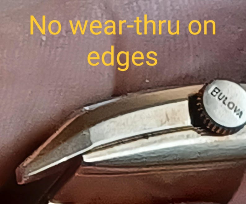K&ouml;nnte beinhalten: Nahaufnahme eines silbernen Uhrenarmbands mit dem Wort "BULOVA" an der Seite. Das Armband weist keine Abnutzung an den Kanten auf. "No wear-thru on edges" ist in gelber Schrift auf braunem Hintergrund geschrieben.