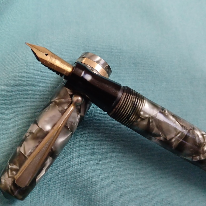 May include: A vintage fountain pen with a gold nib and a marbled gray and black barrel. The pen has a black section and a gold-colored clip. The cap is off, revealing the nib and the pen's internal mechanism.