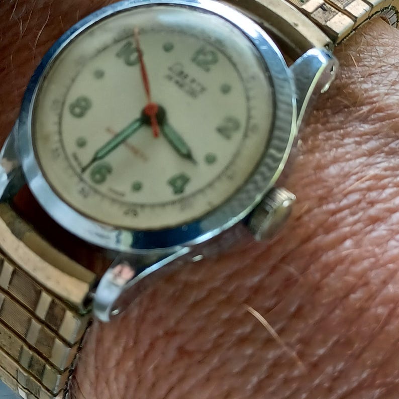 May include: Close-up of a vintage wristwatch with a cream-colored face. The watch features black numerals, a red second hand, and green hour and minute hands. The watch is set in a silver-colored case with a gold-toned metal band.