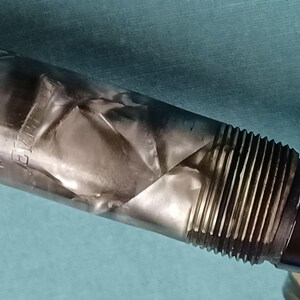 May include: Close-up of a pen with a silver, textured body and a black, ridged grip. The pen's silver body has a unique, folded appearance. The grip section features vertical ridges for enhanced hold.