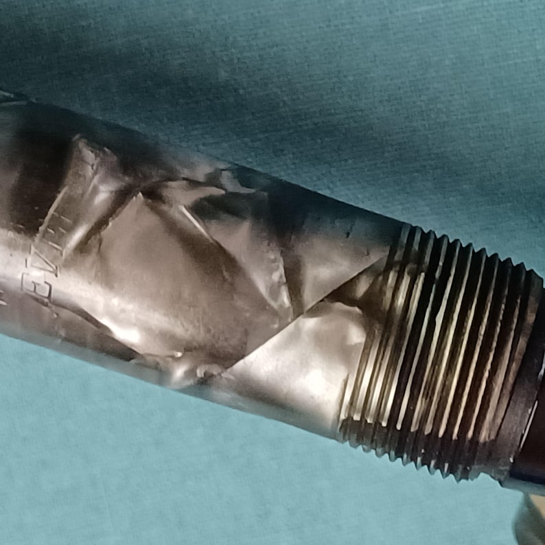 May include: Close-up of a pen with a silver, textured body and a black, ridged grip. The pen's silver body has a unique, folded appearance. The grip section features vertical ridges for enhanced hold.