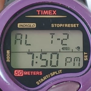 May include: A purple Timex Indiglo digital watch with a black face and white digital display. The watch shows the time as 7:50 PM, the date as T-2, and the alarm is set. The watch is water resistant to 50 meters.