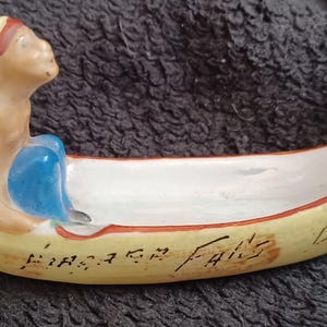 May include: A ceramic canoe ashtray with a painted figure of a Native American paddling. The canoe is painted yellow with red and brown accents. The text "Niagara Falls" is written on the side of the canoe.
