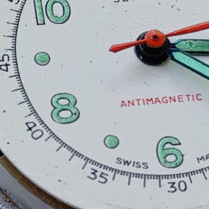 May include: Close-up of a vintage watch face with a white background. The numbers 6, 8, and 10 are in green, with a matching green dot. The words "ANTIMAGNETIC" and "SWISS" are in red and black, respectively. The watch hands are red and blue.