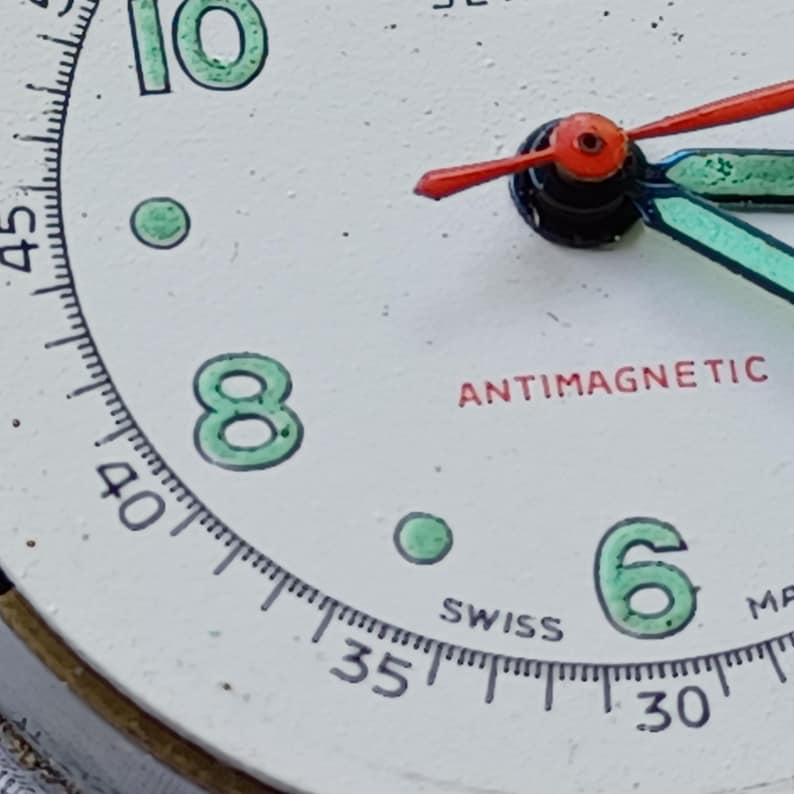 May include: Close-up of a vintage watch face with a white background. The numbers 6, 8, and 10 are in green, with a matching green dot. The words "ANTIMAGNETIC" and "SWISS" are in red and black, respectively. The watch hands are red and blue.
