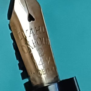 May include: Close-up of a gold-colored fountain pen nib. The nib has the engraved text "WAHL OXFORD 2 14V MADE IN GERMANY". The nib has a heart-shaped breather hole.