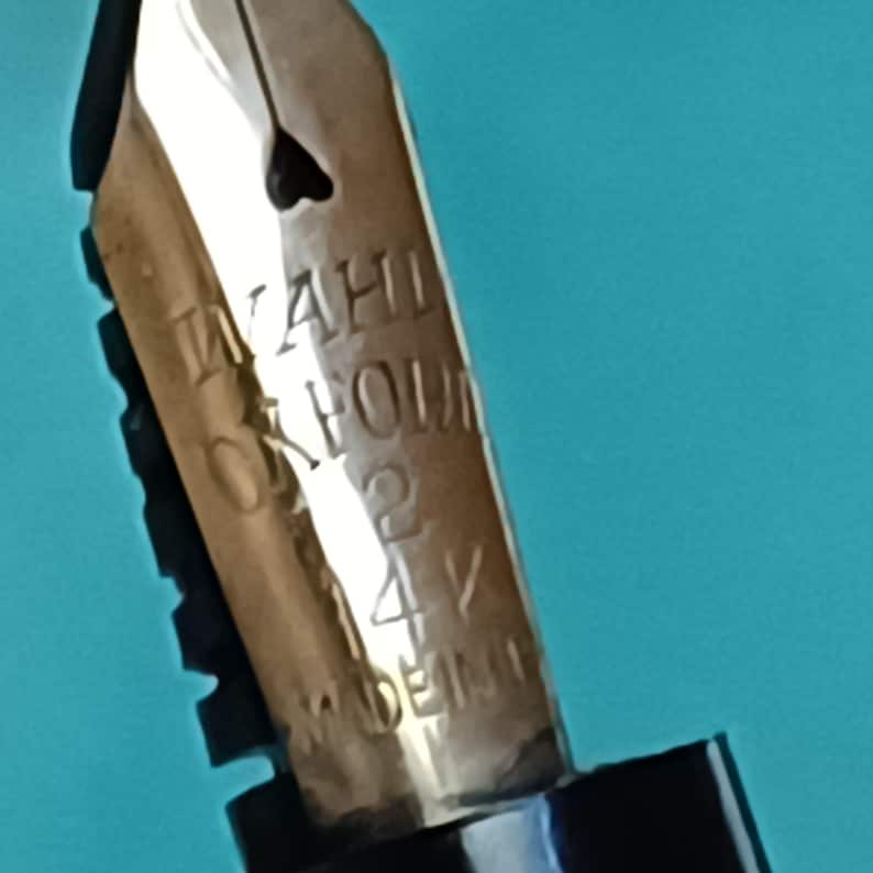 May include: Close-up of a gold-colored fountain pen nib. The nib has the engraved text "WAHL OXFORD 2 14V MADE IN GERMANY". The nib has a heart-shaped breather hole.