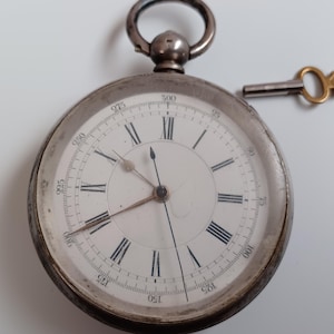 May include: A silver pocket watch with Roman numeral markings on the face and a key attached to the top.