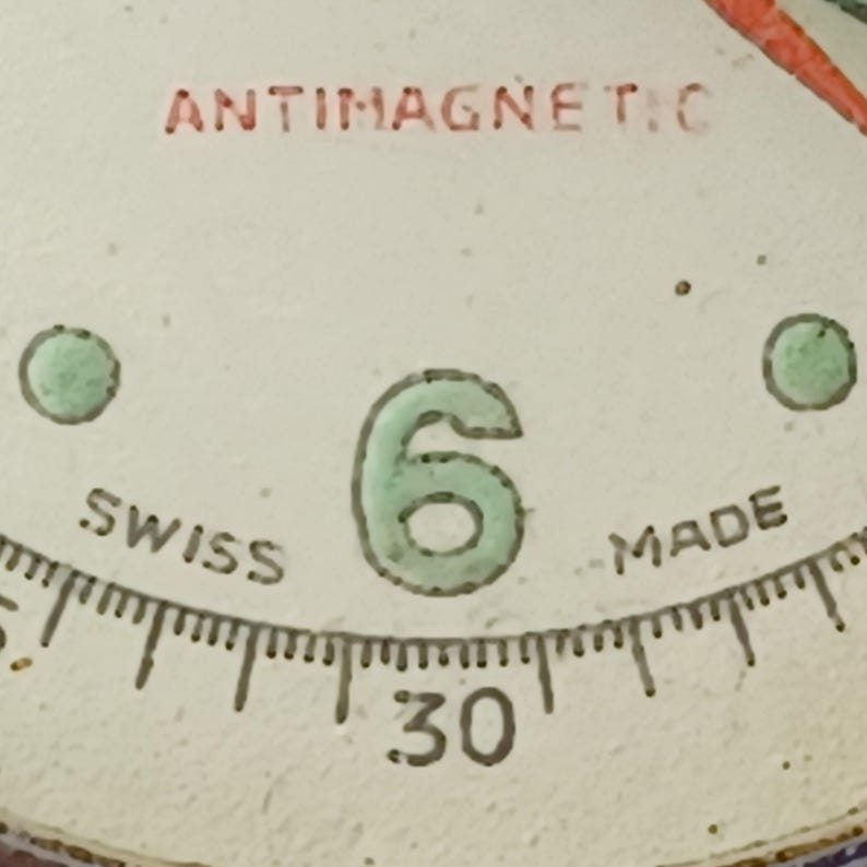 May include: Close-up of a watch face with the word "ANTIMAGNETIC" in red. The number "6" is in green, with "SWISS MADE" and the number "30" visible. The face has a white background with small green dots.