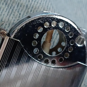 May include: A silver-toned metal lighter with a hinged lid and a textured, striped surface. The top section features a circular opening surrounded by a ring of small, evenly spaced perforations, revealing a central wick inside the mechanism.