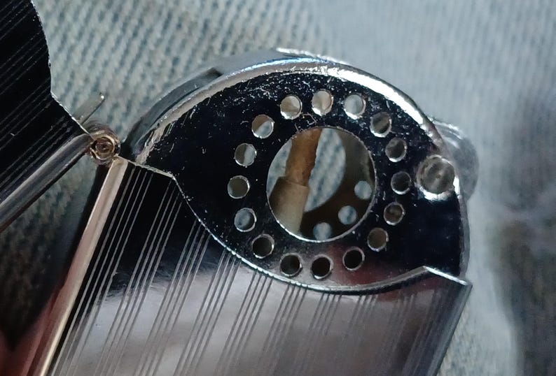 May include: A silver-toned metal lighter with a hinged lid and a textured, striped surface. The top section features a circular opening surrounded by a ring of small, evenly spaced perforations, revealing a central wick inside the mechanism.