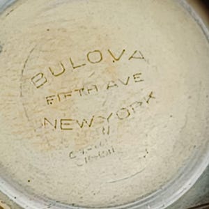 K&ouml;nnte beinhalten: Nahaufnahme eines Zifferblatts mit der Aufschrift "BULOVA FIFTH AVE NEW YORK" in einem kreisf&ouml;rmigen Design.