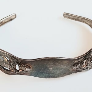 May include: A silver bracelet with an ornate design. The bracelet is made of a single piece of metal that is bent into a circle. The design features a floral pattern and a smooth, flat center section.