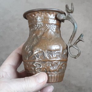 May include: A small, copper pitcher with an ornate design featuring animals and floral motifs. The pitcher has a hinged lid and a handle with a decorative finial.