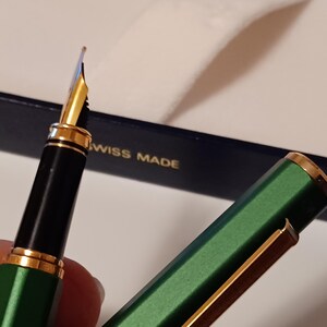 Puede incluir: Una pluma estilogr&aacute;fica verde y dorada con un cartucho de tinta negro y una plumilla de oro. La pluma est&aacute; en una caja azul con las palabras "Swiss Made" impresas en ella.