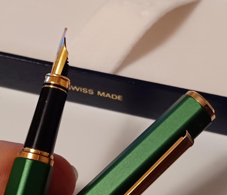 Puede incluir: Una pluma estilogr&aacute;fica verde y dorada con un cartucho de tinta negro y una plumilla de oro. La pluma est&aacute; en una caja azul con las palabras "Swiss Made" impresas en ella.