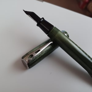May include: A vintage fountain pen with a dark green marbled barrel and cap. The pen features a black nib and a silver-colored clip. The cap is partially removed, revealing the pen's writing tip. The pen is a classic writing instrument.