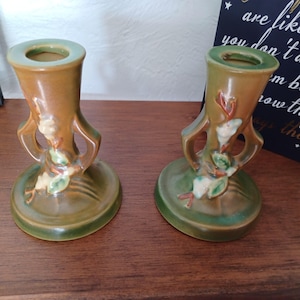 May include: A pair of vintage green and brown ceramic candle holders with floral designs. The holders have a unique shape with two handles on each side.
