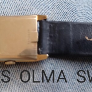 K&ouml;nnte beinhalten: Eine goldfarbene Armbanduhr mit einem schwarzen Lederband. Das Band ist mit den Worten "OLMA SWISS" gepr&auml;gt.