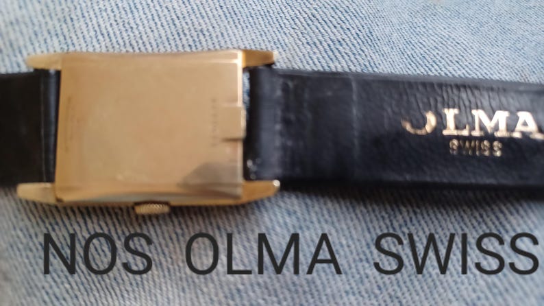 K&ouml;nnte beinhalten: Eine goldfarbene Armbanduhr mit einem schwarzen Lederband. Das Band ist mit den Worten "OLMA SWISS" gepr&auml;gt.